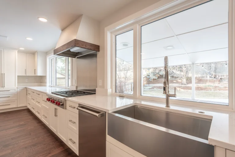 Bright modern kitchen with white cabinets, stainless steel appliances, and large windows overlooking a backyard garden