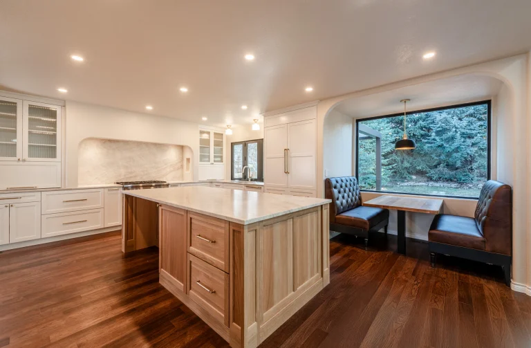 Bright modern kitchen with white cabinets, marble backsplash, a wooden island, and a windowed dining nook overlooking trees.