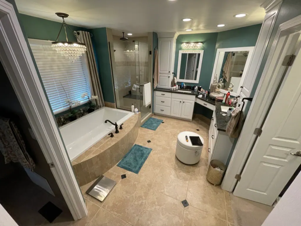 Spacious bathroom with a corner bathtub, glass shower, and white cabinetry against teal walls; dual vanities and a round ottoman in the center.