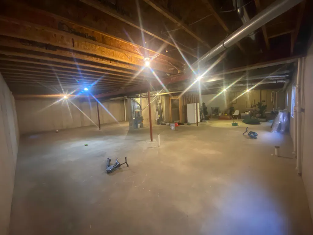 Unfinished basement with exposed ceiling beams, concrete floor, and scattered miscellaneous items under bright lights.
