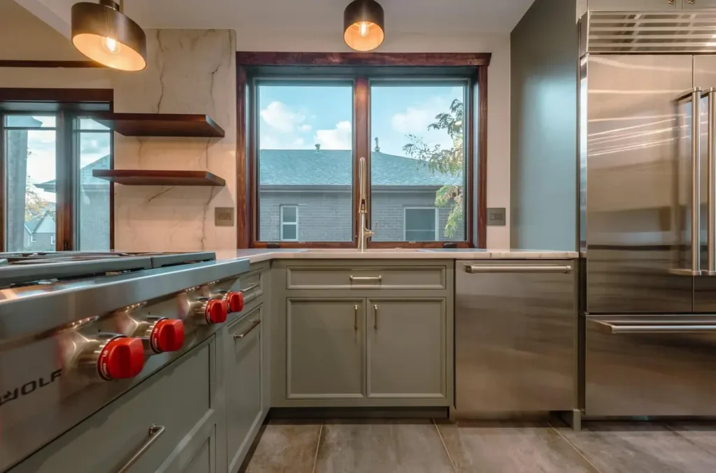 Sage green kitchen cabinets around a pro range with red knobs and a stainless refrigerator